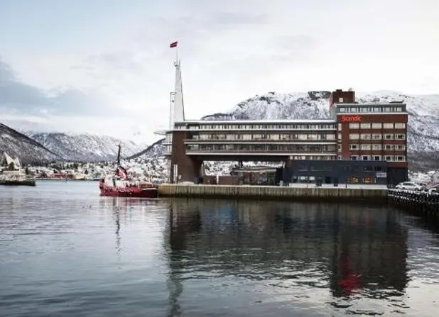 A waterfront building with a red Scandic sign and a tall mast stands on a pier in front of snow-covered mountains.